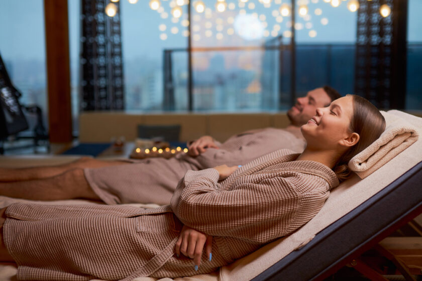 A man and woman relax on loungers in a spa, wearing matching brown robes. The room is softly lit with candles, and large windows show city lights in the background. They appear serene and comfortable.