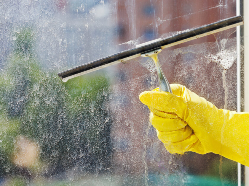 A gloved hand uses a squeegee to clean a dirty window. Soap suds can be seen on the glass. The blurred background shows an outdoor scene with a building and greenery.