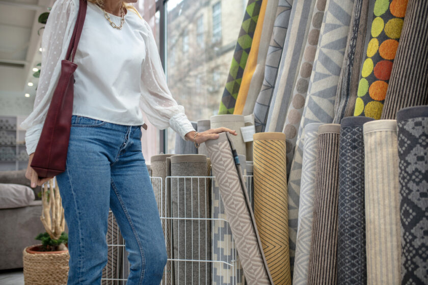 Carpets, assortment. Woman in a white blouse and jeans standing in the carpet department of a furniture store, holding a rug with her hand, her face is not visible.