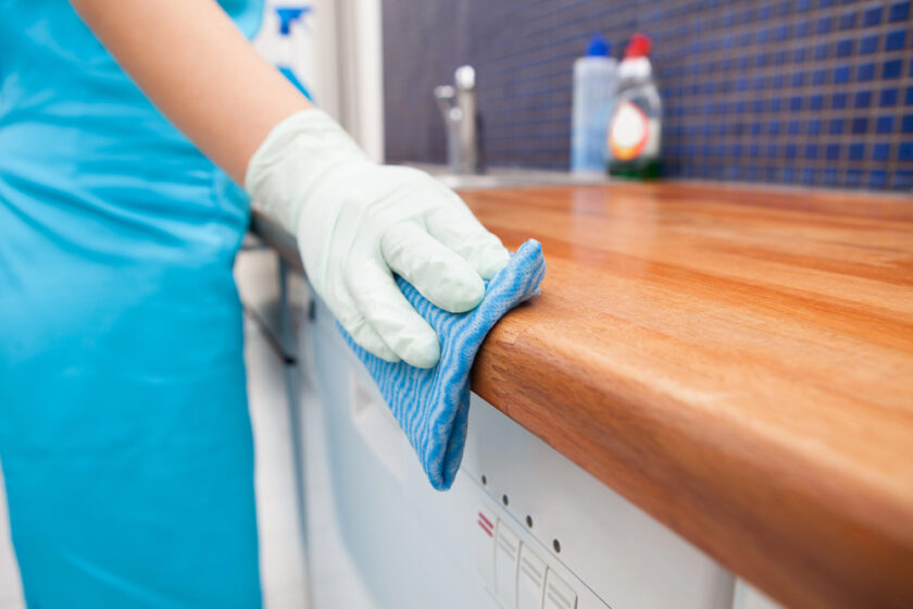A person wearing a blue apron and white gloves cleans a wooden kitchen counter with a blue cloth. In the background, there are cleaning supplies on the counter near a sink with blue tiled walls.