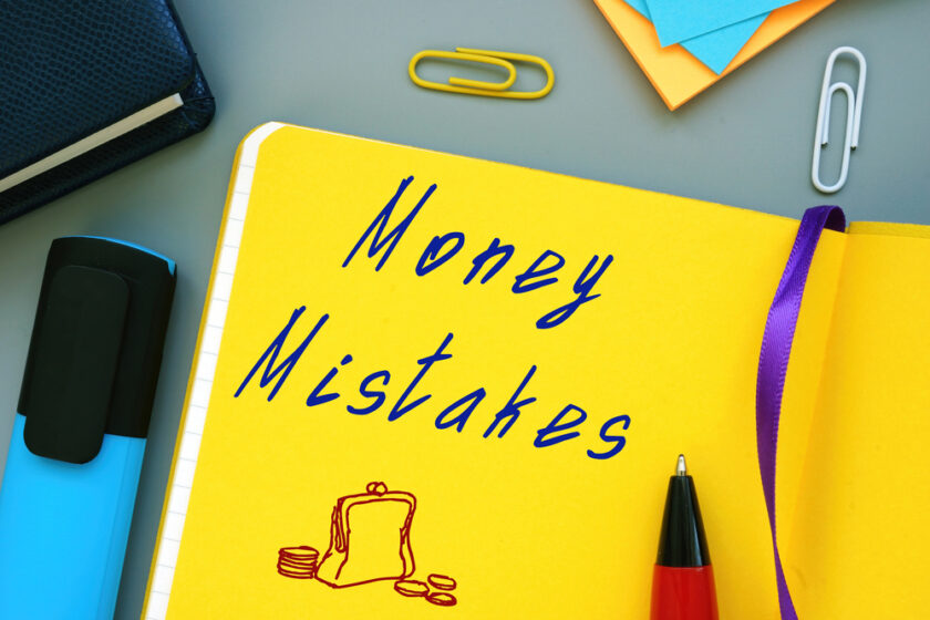 A yellow notebook with Money Mistakes written in blue marker is open on a desk. Beside it are a black pen, a blue highlighter, colored paper clips, and sheets of paper. A drawing of a wallet and coins is visible.