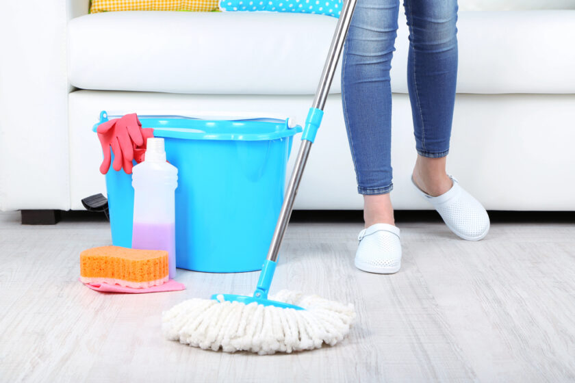 A person wearing jeans and white slippers is cleaning a light wooden floor with a mop. A blue bucket, pink rubber gloves, a sponge, and a cleaning bottle are on the floor nearby, next to a white sofa.