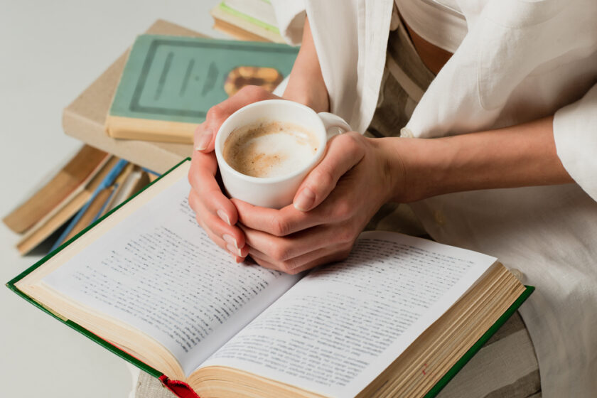 A person in a white shirt holds a cup of frothy coffee over an open book. Several other books are stacked nearby, with a beige surface beneath. The scene suggests a cozy reading and coffee-drinking moment.