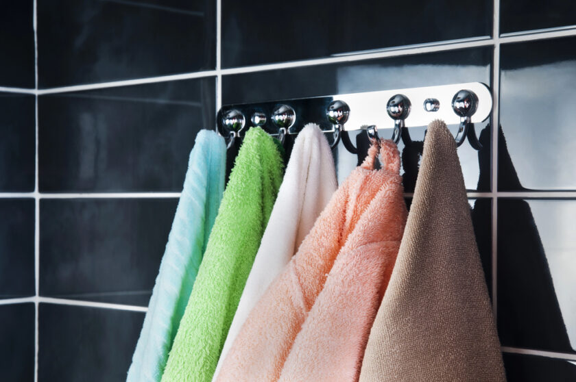 Four towelling robes in blue, green, pink, and beige hang on hooks against a glossy black tiled wall.