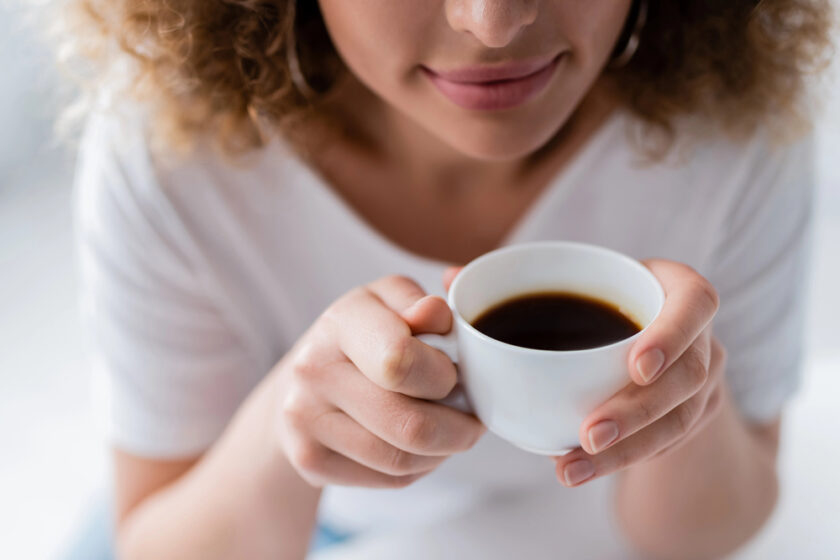 A person with curly hair holds a white cup of black coffee close to their face. They are wearing a white shirt and have neutral-colored nail polish. The focus is on the cup and hands, with soft lighting in the background.