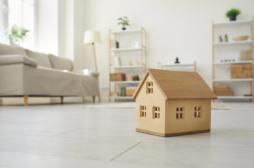 A small wooden house model sits on a light-colored wooden floor in a modern living room. In the background, theres a beige sofa, a tall lamp, and shelves with books and decor items near a window.