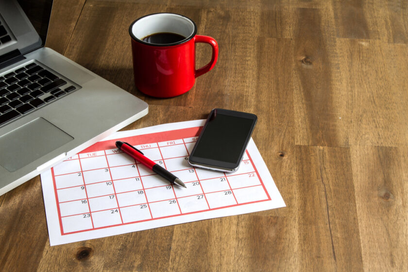 A red coffee mug, a smartphone, and a pen are placed on a calendar on a wooden table next to a laptop. The calendar displays a week, with days Tuesday to Friday visible.