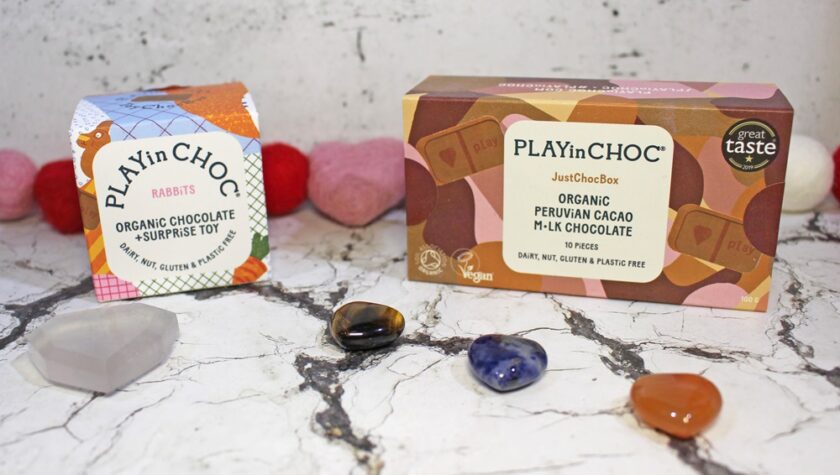 Two boxes of PLAYin CHOC organic chocolate are on a marble surface with four polished stones in front and felt heart decorations in the background. The left box is for rabbits, the right is Peruvian cacao milk chocolate.