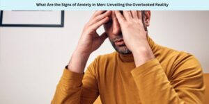 A man in a mustard turtleneck sits with eyes closed, massaging his temples with his fingers. A headline above reads, What Are the Signs of Anxiety in Men: Unveiling the Overlooked Reality.