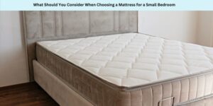 A neatly made bed with a thick, white quilted mattress on a beige upholstered bed frame. The room has wooden flooring and neutral-toned walls. Above the bed, text reads: What Should You Consider When Choosing a Mattress for a Small Bedroom.