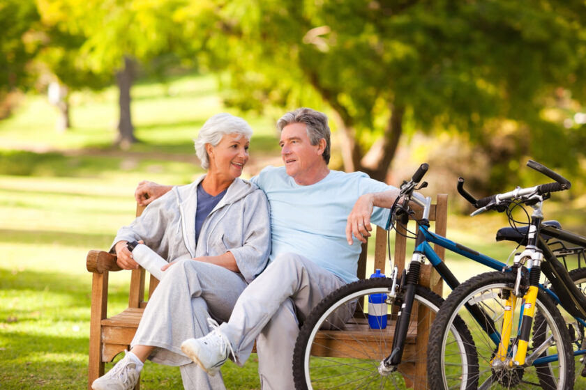 An elderly couple sits on a park bench, smiling at each other. They are dressed in casual clothing, and two bicycles are parked nearby. The background is a lush green park with trees, suggesting a pleasant, sunny day.