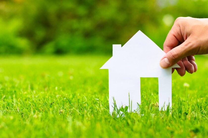 A hand holds a white paper cutout of a house against a backdrop of vibrant green grass, symbolizing eco-friendly living or real estate concepts. The background is blurred, highlighting the house cutout in the foreground.
