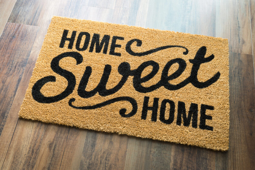 A coir doormat on a wooden floor with the words Home Sweet Home printed in bold black lettering. The word Sweet is stylized in cursive.