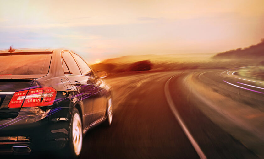 A sleek black car speeds down a winding road at sunset. The background is blurred, showcasing motion and a vibrant sky with warm orange and pink hues.