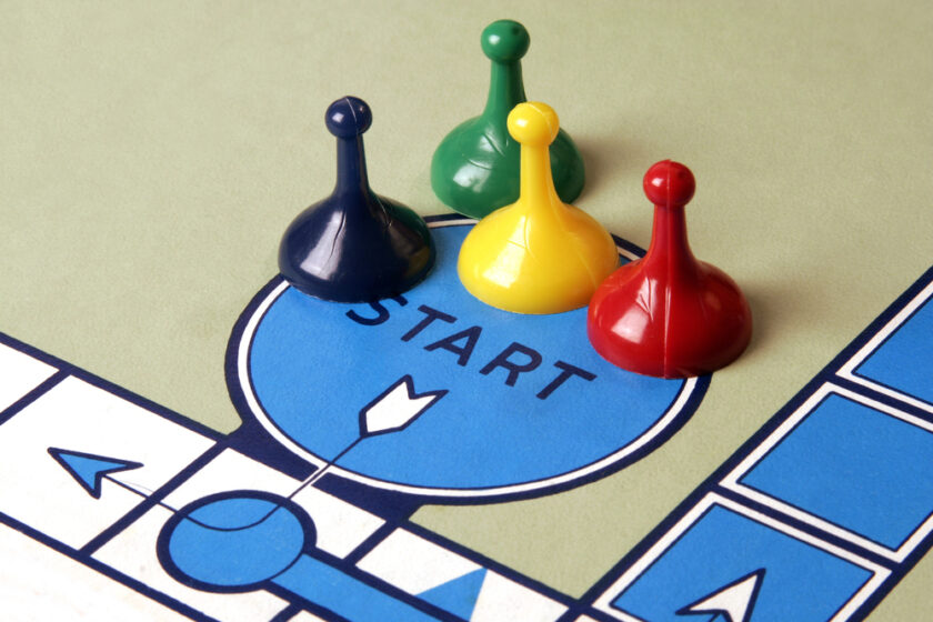 Four colorful game pieces—blue, yellow, green, and red—are placed on a board game start space, each shaped like a classic pawn. The board features a blue start circle and paths marked with arrows.