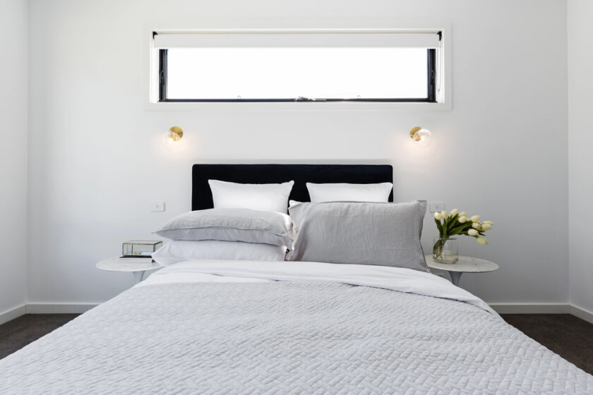 A minimalist bedroom featuring a neatly made bed with gray and white bedding. Two small round nightstands flank the bed, each with a white lamp and a bouquet of yellow tulips. A rectangular window above the bed brightens the room.