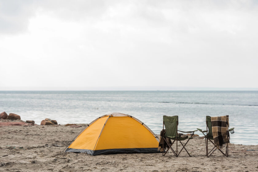 A yellow tent and two folding chairs with a blanket sit on a sandy beach by a calm sea under a cloudy sky.