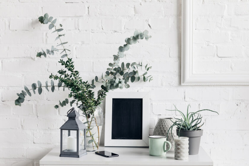 A white table against a textured white brick wall holds greenery in vases, a small framed chalkboard, a decorative lantern with a candle, a smartphone, a mug, and a potted plant, creating a minimalist and cozy decor scene.