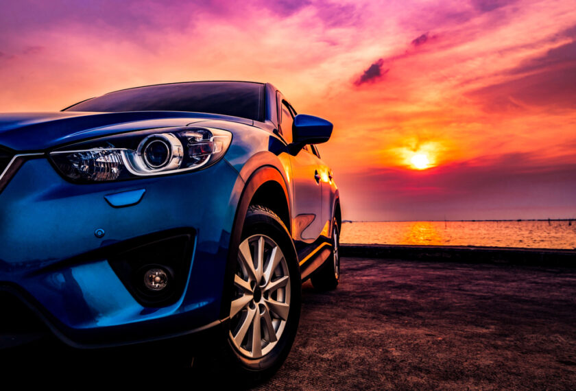 Blue SUV parked on a concrete surface, with a vibrant sunset over the ocean in the background. The sky is filled with orange, pink, and purple hues, and the sun is low near the horizon. The vehicles front-left side is prominently displayed.