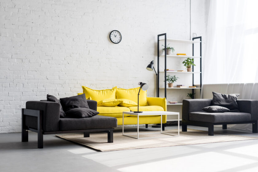 A modern living room with a bright yellow sofa, two dark gray armchairs, and a white coffee table on a beige rug. A black floor lamp stands nearby, and a white bookshelf with plants is against a white brick wall. A small clock hangs above.