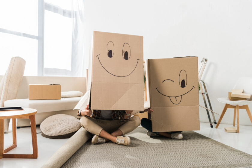 Two people sit on the floor in a bright room, each with a large cardboard box over their heads. The boxes have smiley face drawings; one winks with its tongue out. Around them are more boxes, a chair, and soft furnishings.