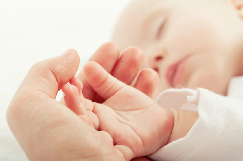 Close-up of an adults hand gently holding a sleeping babys hand. The image conveys tenderness and care, focusing on the delicate fingers and soft skin. The background is softly blurred to emphasize the connection.