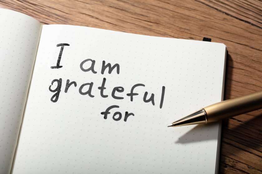 Open notebook on a wooden surface with the words I am grateful for written in black ink. A gold pen is placed diagonally across the bottom right corner of the page.