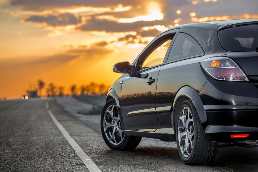 A black car is parked on the side of a road during a sunset. The sky is filled with orange and yellow hues, and the sun is partially obscured by clouds in the background. The road is empty, stretching into the distance.