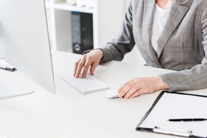 Person in a gray plaid suit working on a computer at a white desk. One hand is on a mouse and the other on a keyboard. A clipboard with a pen is on the desk, and shelves with books are visible in the background.