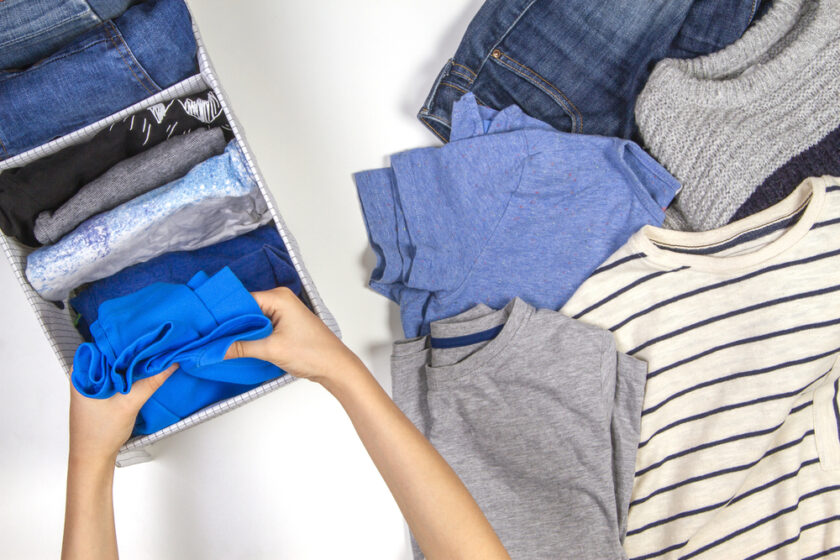 Hands folding blue clothing next to a drawer with neatly organized clothes. Various tops, including a striped shirt and gray t-shirt, are spread around. A pair of jeans and a gray sweater are partially visible.