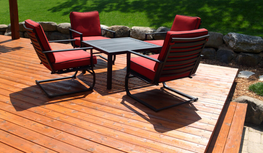 Four red-cushioned patio chairs surround a rectangular black table on a wooden deck. The deck overlooks a green lawn bordered by rocks. Sunlight casts shadows on the decks surface.