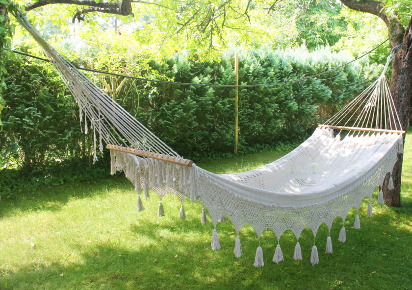 A white, woven hammock hangs between two trees in a sunny, grassy garden. The hammock features intricate patterns and fringe tassels. Lush green hedges and foliage surround the peaceful scene.