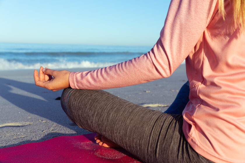 A person in a pink long-sleeve shirt and gray leggings is sitting cross-legged on a red mat at the beach, facing the ocean. They are meditating with their hand resting on their knee, and the sun is shining on the waves.