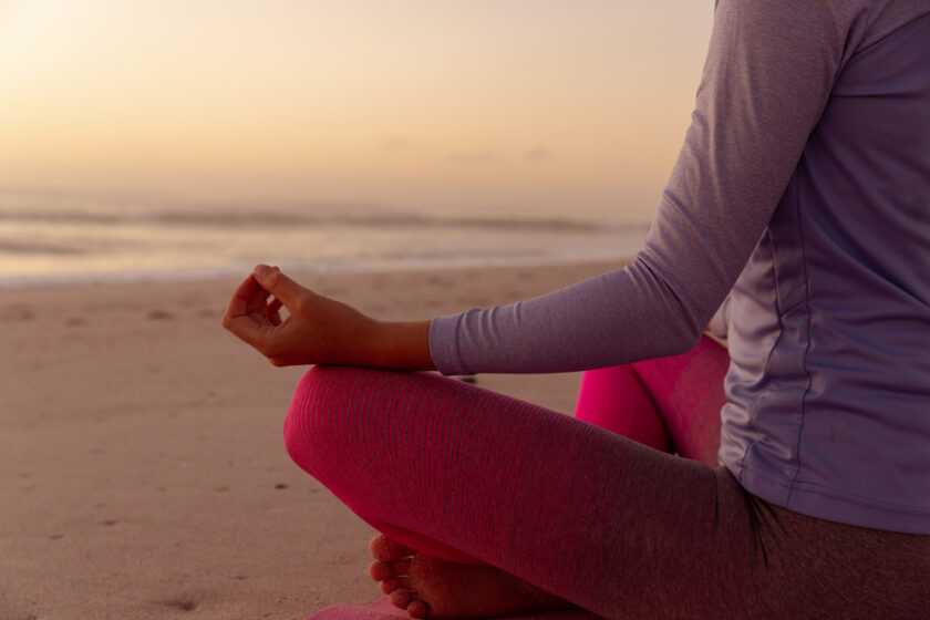 Person seated in a cross-legged position on a beach, meditating at sunset. They are wearing a light gray long-sleeve shirt and pink leggings. The ocean and a warm, glowing sky with soft clouds are in the background.