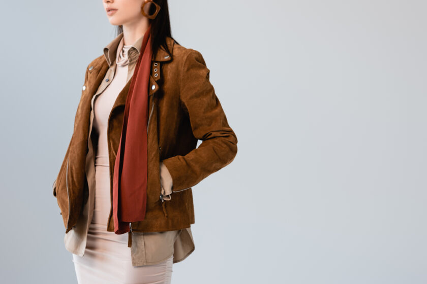 A person wearing a brown leather jacket over a beige shirt and skirt, accessorized with a red scarf and large round earrings, stands against a simple light background. Their hands are in the jacket pockets, and their face is not visible.