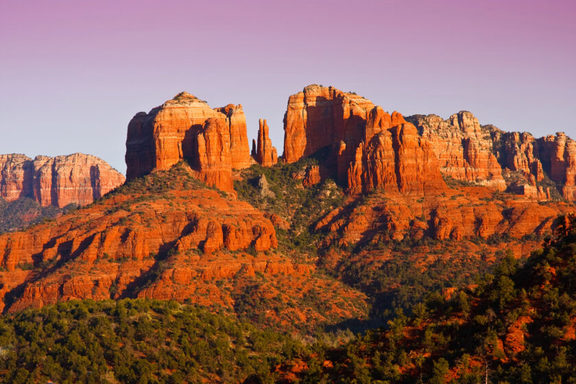 Red rock formations stand tall under a pink and purple sky at sunset in Sedona, Arizona. The layered cliffs and mesas are surrounded by green vegetation, creating a striking contrast between the red rocks and the vibrant sky.