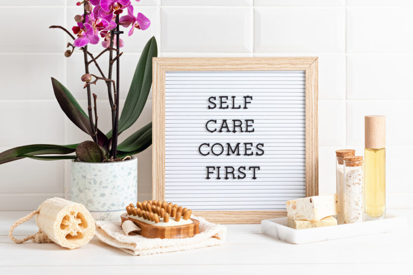 A framed board with the text Self Care Comes First is placed on a white tile surface. Nearby are a potted orchid, a loofah, a wooden massage tool, soaps, and a bottle of oil, creating a relaxing atmosphere.