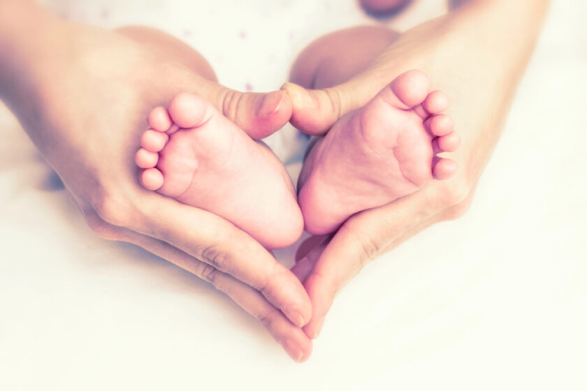 Adult hands gently cradle baby feet in a heart shape, set against a soft, blurred background. The image conveys warmth and tenderness.