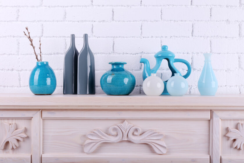 Ceramic vases and decorative bottles in shades of blue and gray are arranged on a light wooden mantel with ornate carvings. The background features a white brick wall.