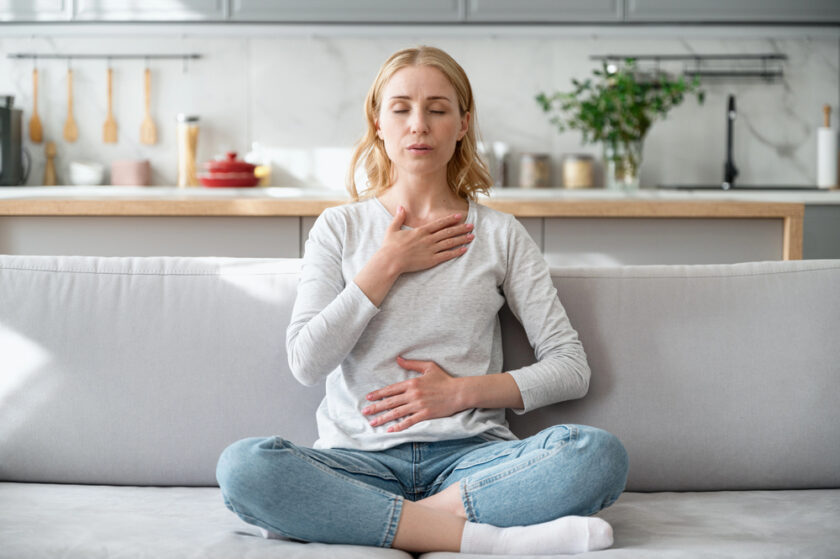 A woman sits cross-legged on a couch in a modern kitchen, with eyes closed. She places one hand on her chest and the other on her stomach, practicing deep breathing or meditation. She wears a light gray sweater and jeans.