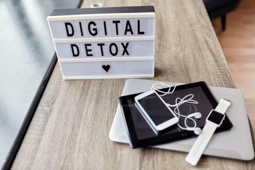 A lightbox sign reading DIGITAL DETOX with a heart below is placed on a wooden table. Nearby are a smartphone, a tablet, earphones, and a smartwatch stacked on a closed laptop.