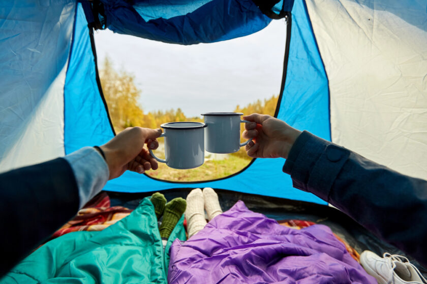Inside a tent, two people clink metal mugs over sleeping bags. Their legs are seen in colorful socks. The tent opens to a view of grass and autumn trees, creating a cozy camping atmosphere.