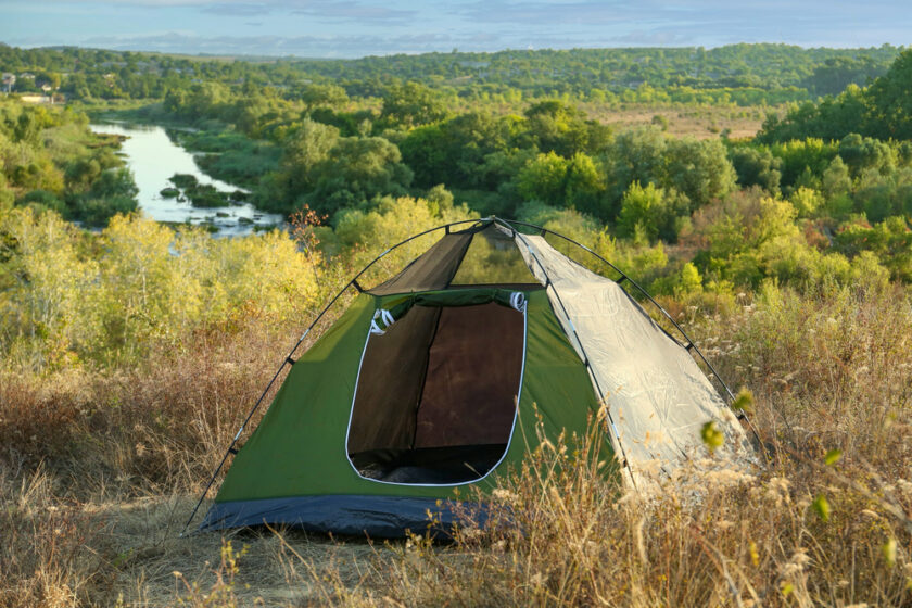 A green camping tent on a dry, grassy hill overlooking a scenic river. The background features lush trees and rolling hills under a partly cloudy sky.