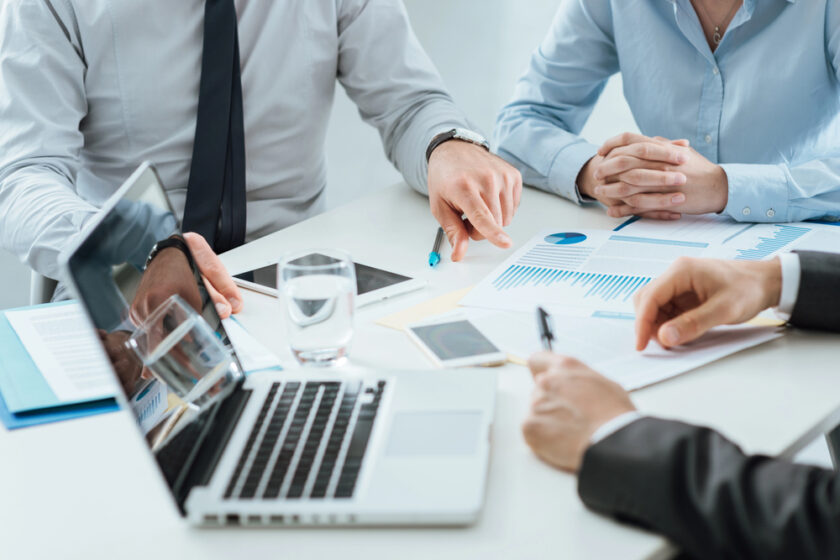 Three individuals in business attire are sitting around a table with a laptop, documents with charts, a tablet, and a glass of water. One person gestures towards a document while the others listen attentively.