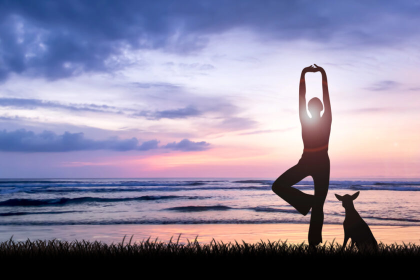 Silhouette of a person practicing yoga in a tree pose, standing on one leg with arms raised, on a beach at sunset. A dog sits nearby, with the ocean and colorful sky in the background.