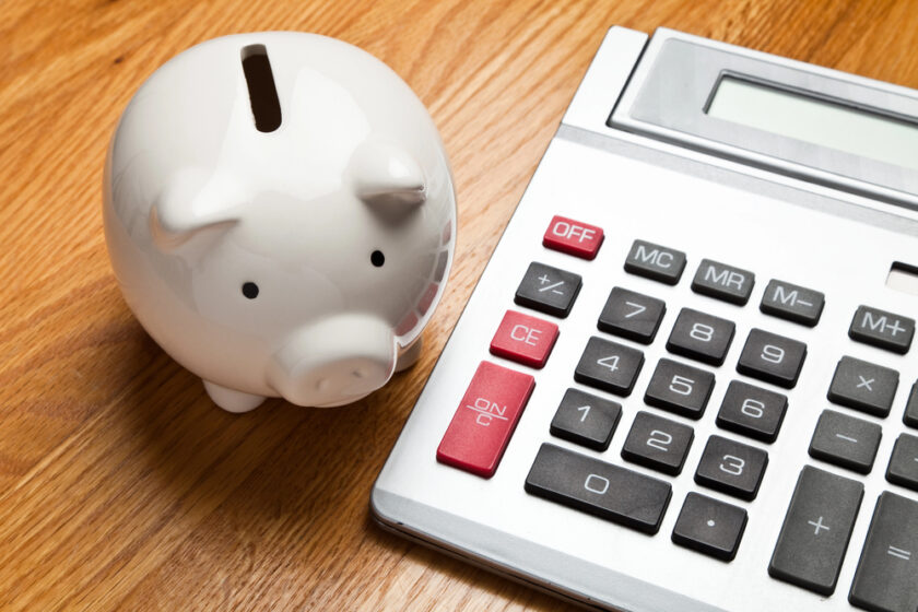 A white ceramic piggy bank is placed on a wooden surface beside a large calculator with red and black buttons. The calculators display screen is visible, and both items suggest themes of savings and financial management.