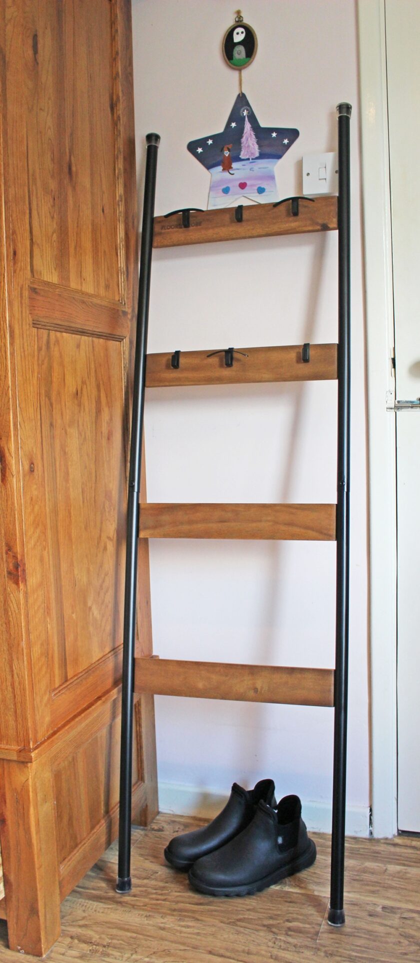 A wooden and metal ladder with four hooks leans against a wall next to a wooden cabinet. A pair of black boots sits on the floor beneath the ladder. A colorful drawing is displayed on the top rung.
