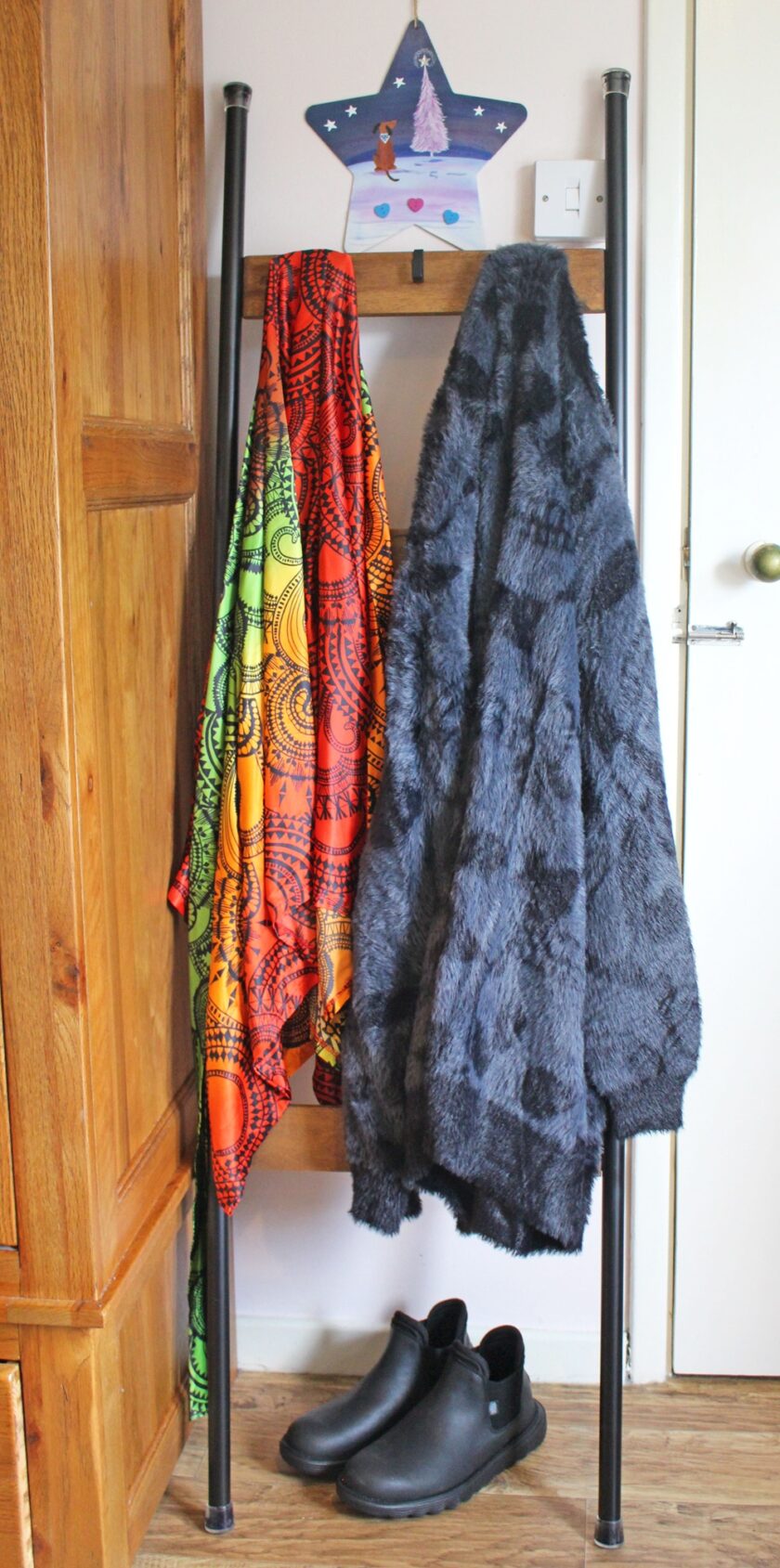 The floordrobe holds a vibrant, multicolored scarf and a fuzzy, dark gray jacket. Below, a pair of black shoes sits on the floor. A star-shaped wall decoration featuring a castle and reindeer hangs above.