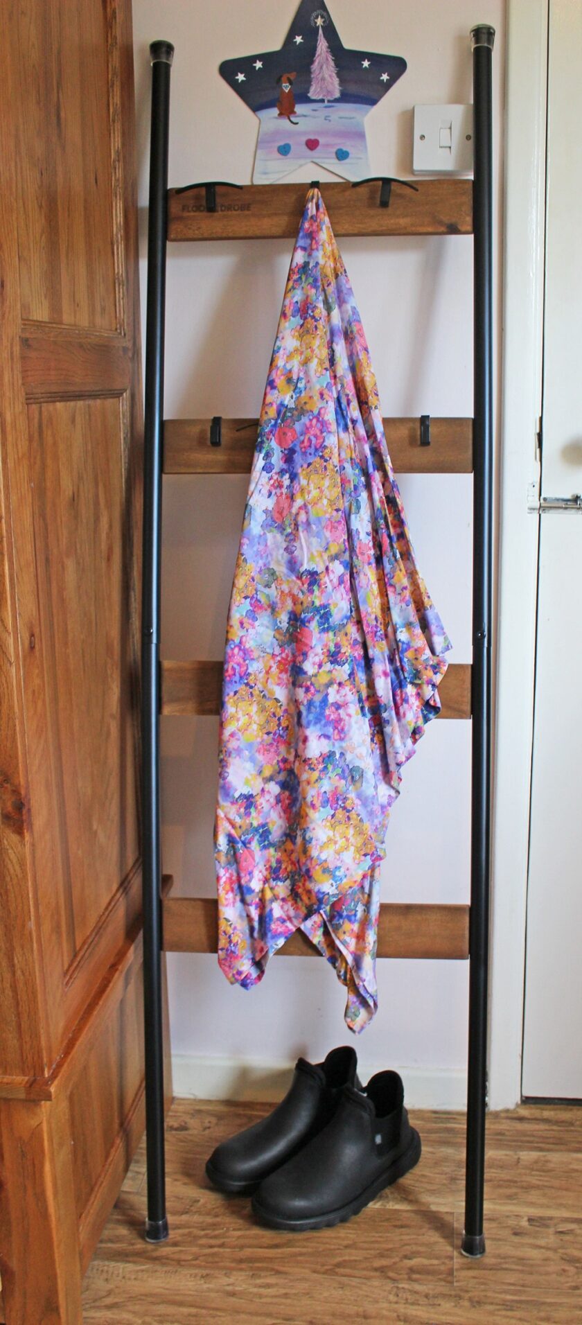 A colorful floral-patterned fabric drapes over a black and wooden ladder propped against a wall in a room. Black shoes are placed on the floor below the ladder. A wooden star with a princess illustration is at the top of the ladder.
