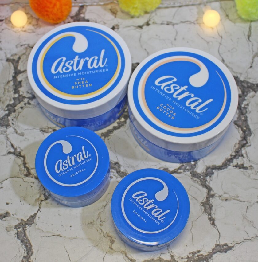 Four blue containers of Astral moisturizer are displayed on a marble surface. Two larger tubs are labeled Intensive Moisturiser with Shea Butter and with Cocoa Butter, and two smaller ones are marked Original. Multicolored pom-poms are in the background.